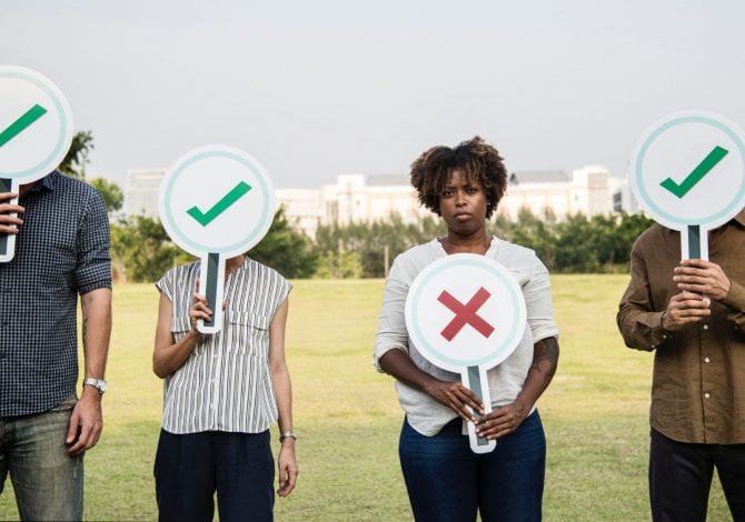 Student Services - Four people standing in a line, holding magnifying glass shaped signs in front of their faces, three have ticks on and one has a cross on, the cross is held below the persons face and she looks sad.