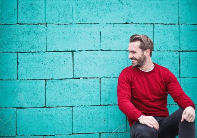 Student Services - Smiling man in red jumper crouched leaning against teal coloured wall.